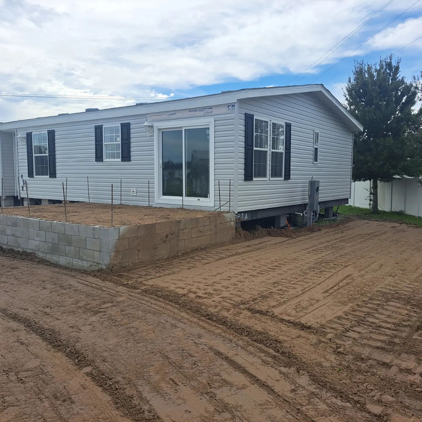 A white mobile home with black shutters is sitting on top of a dirt field.