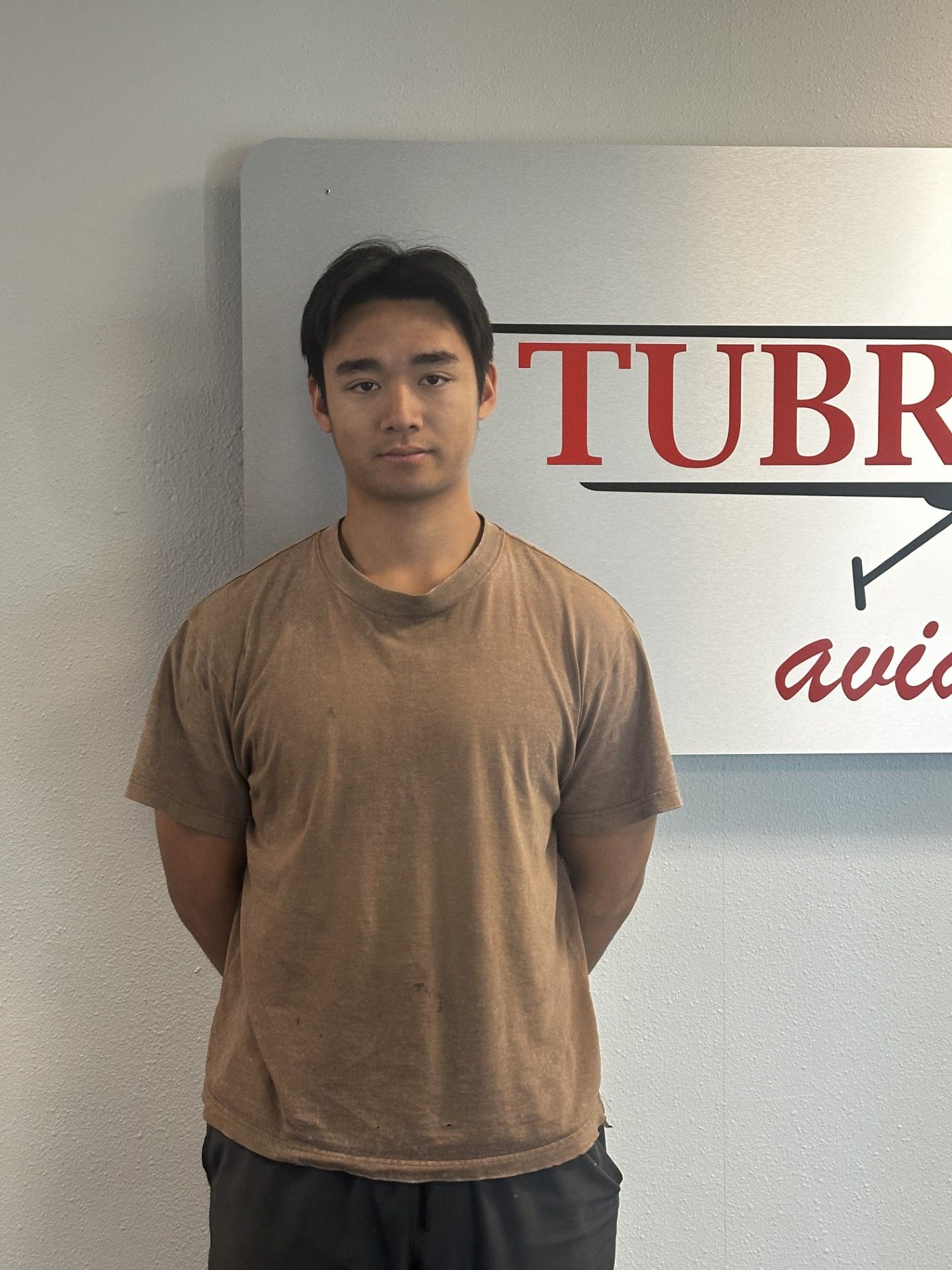 Person standing indoors in front of a sign, wearing a brown T-shirt.