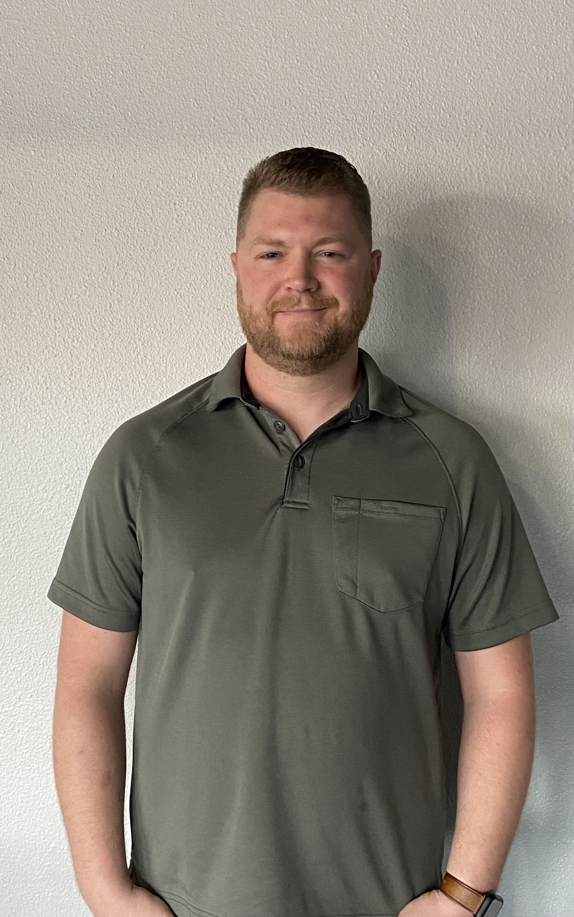 Man in gray polo shirt standing against a light wall, looking at the camera.