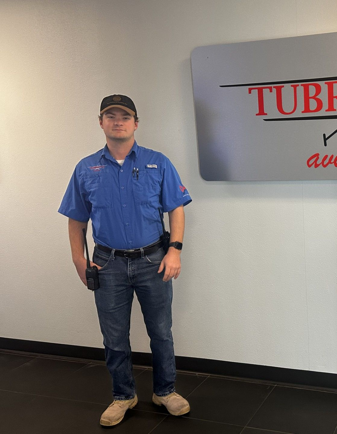 Man in blue work shirt and jeans standing beside a wall sign indoors.
