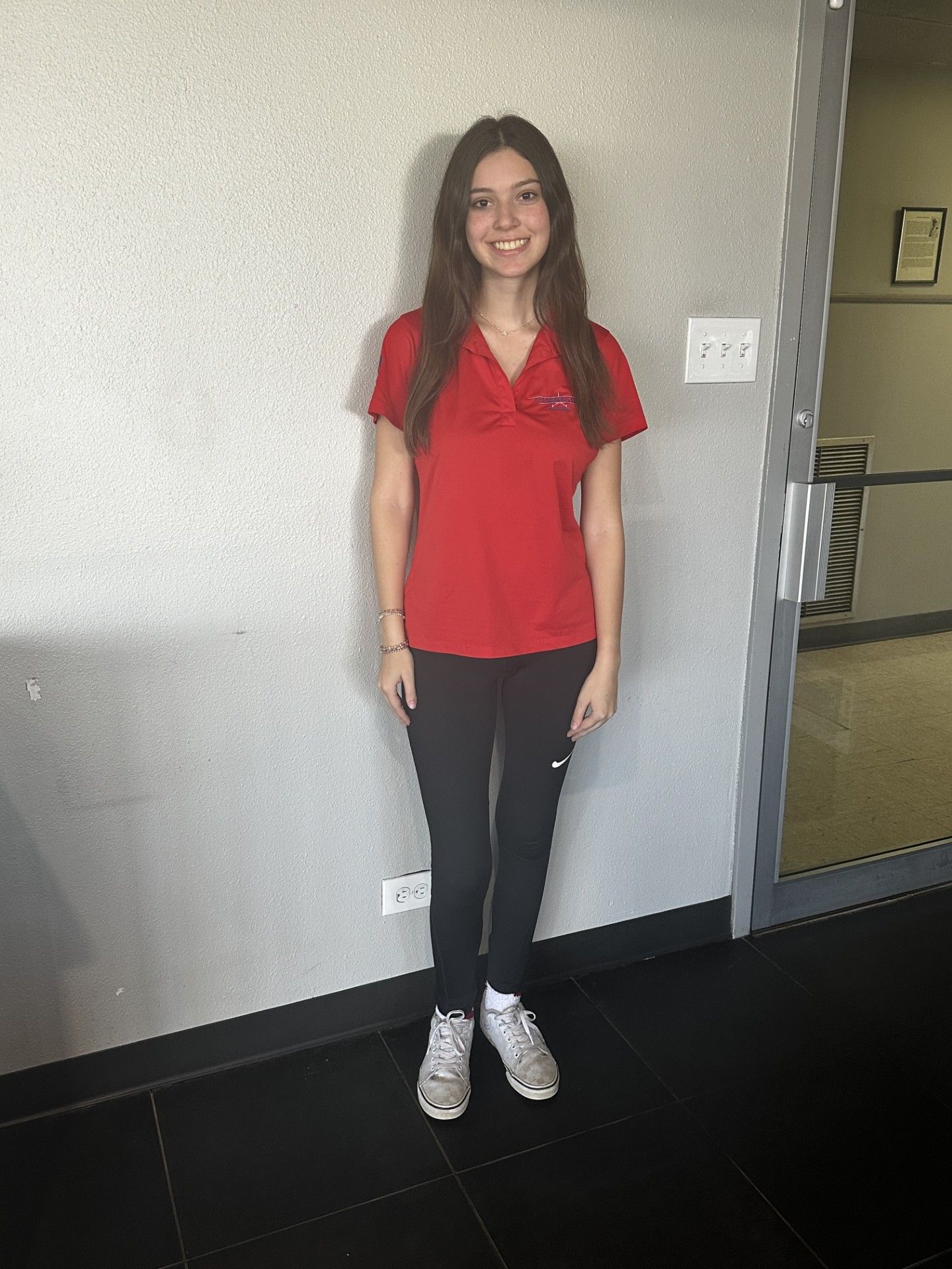Woman in red shirt and black pants standing indoors against a wall, smiling at the camera.
