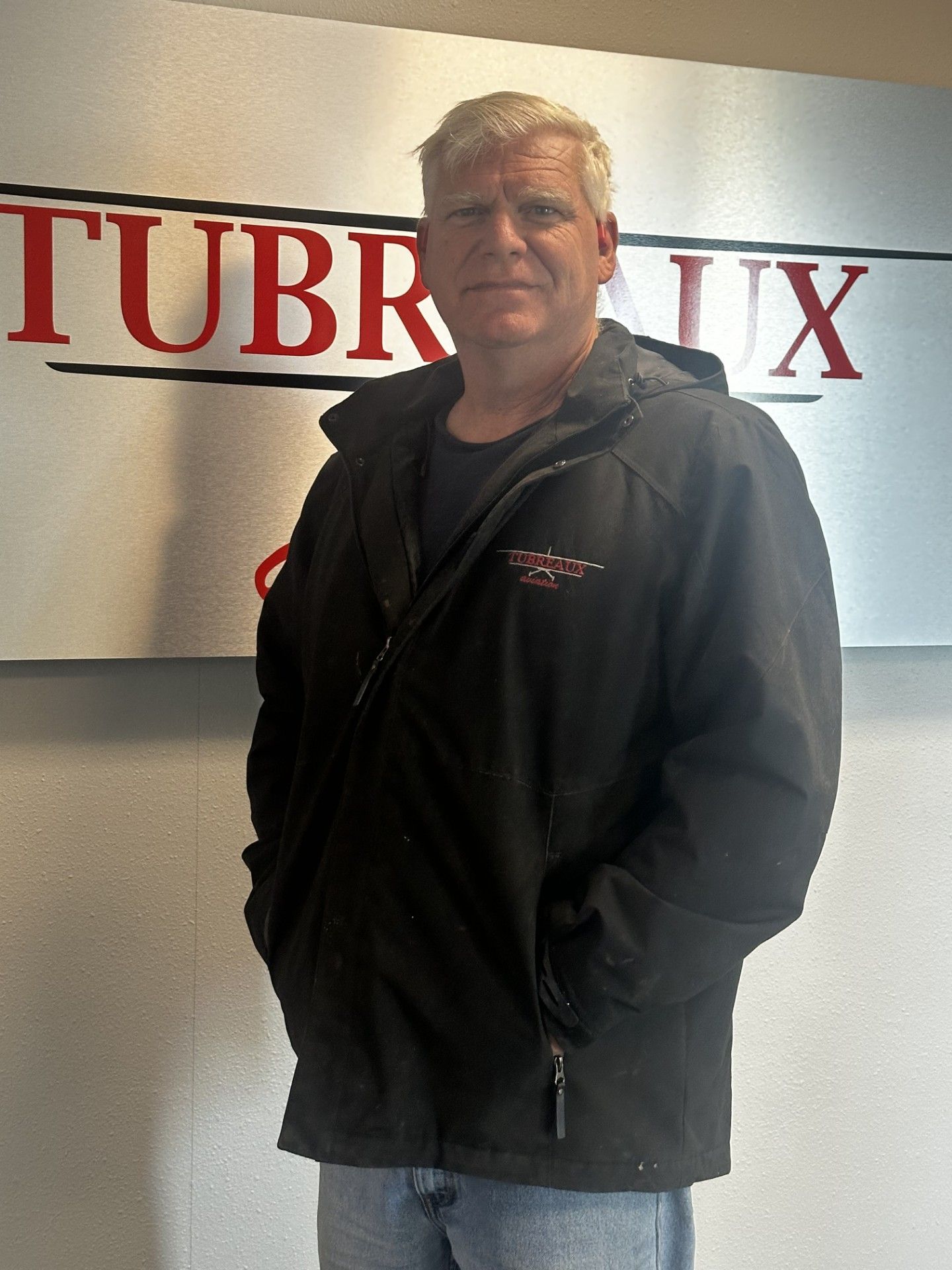 Man standing indoors in a black jacket in front of a white wall with red lettering