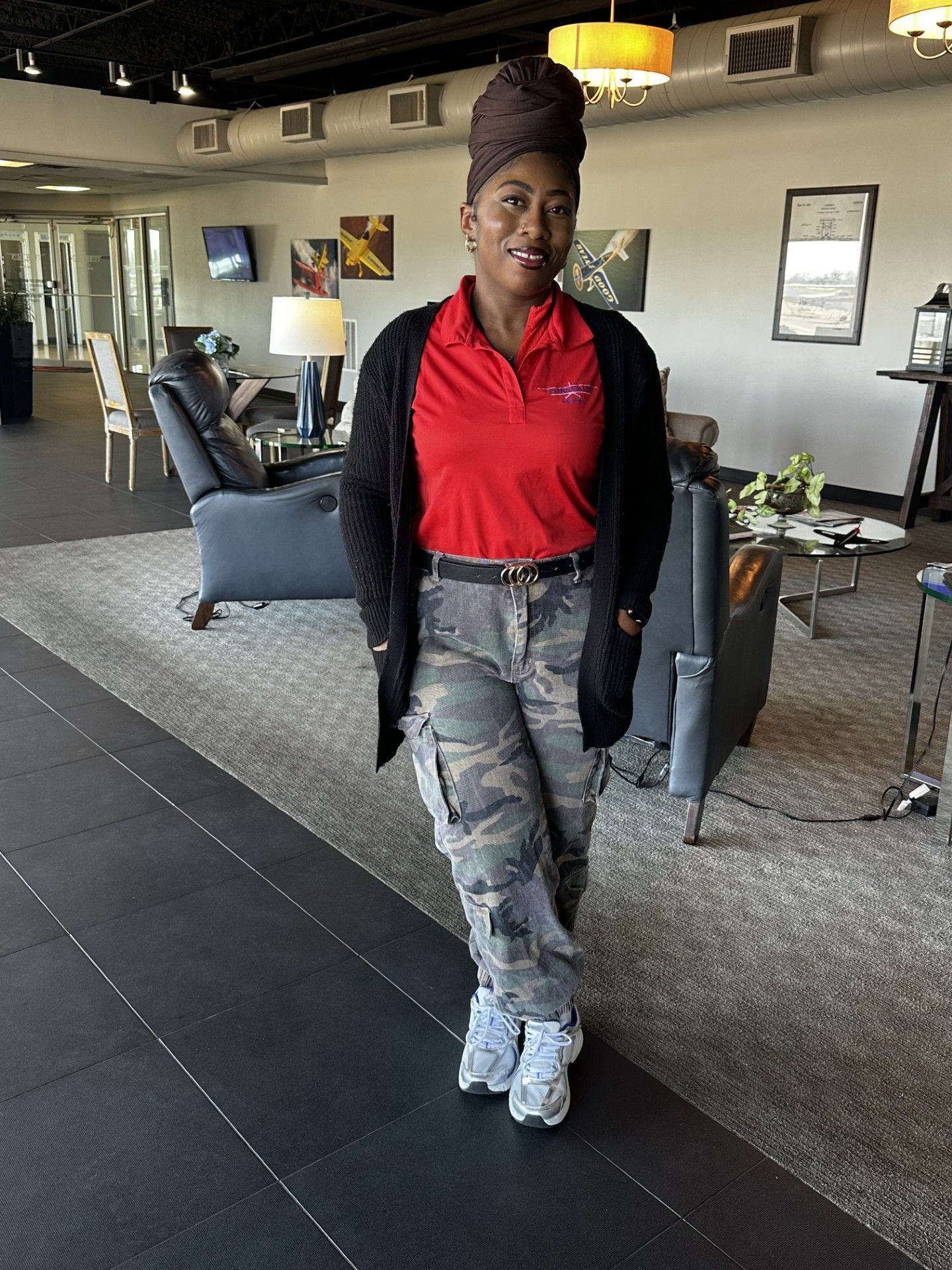 Person standing in a lounge, wearing a red shirt, black cardigan, gray jeans, and white boots.