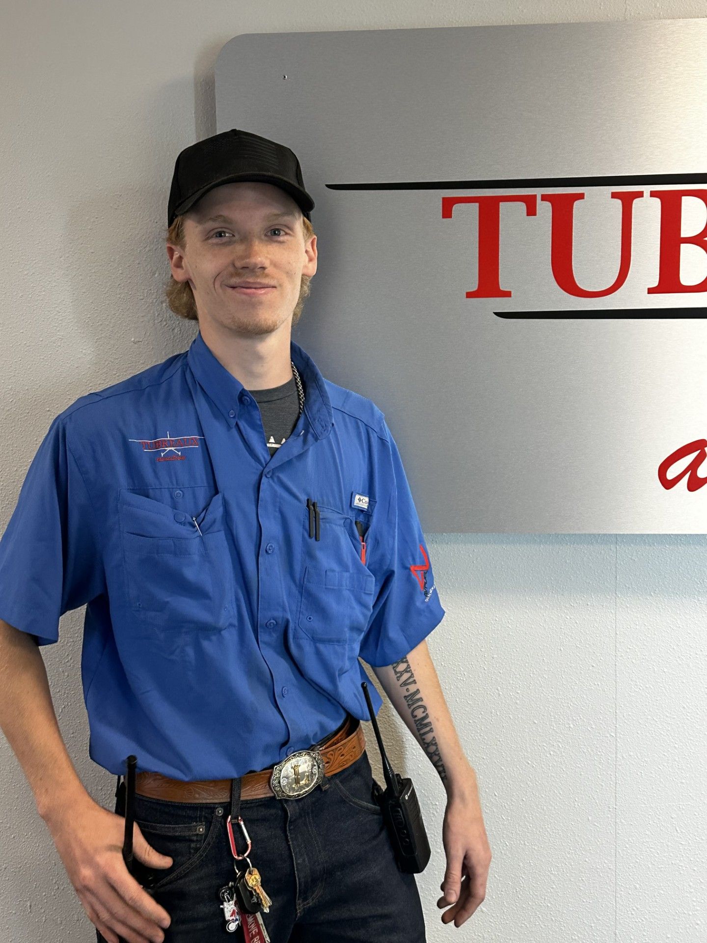 Man in blue work shirt and black cap stands beside a sign indoors.