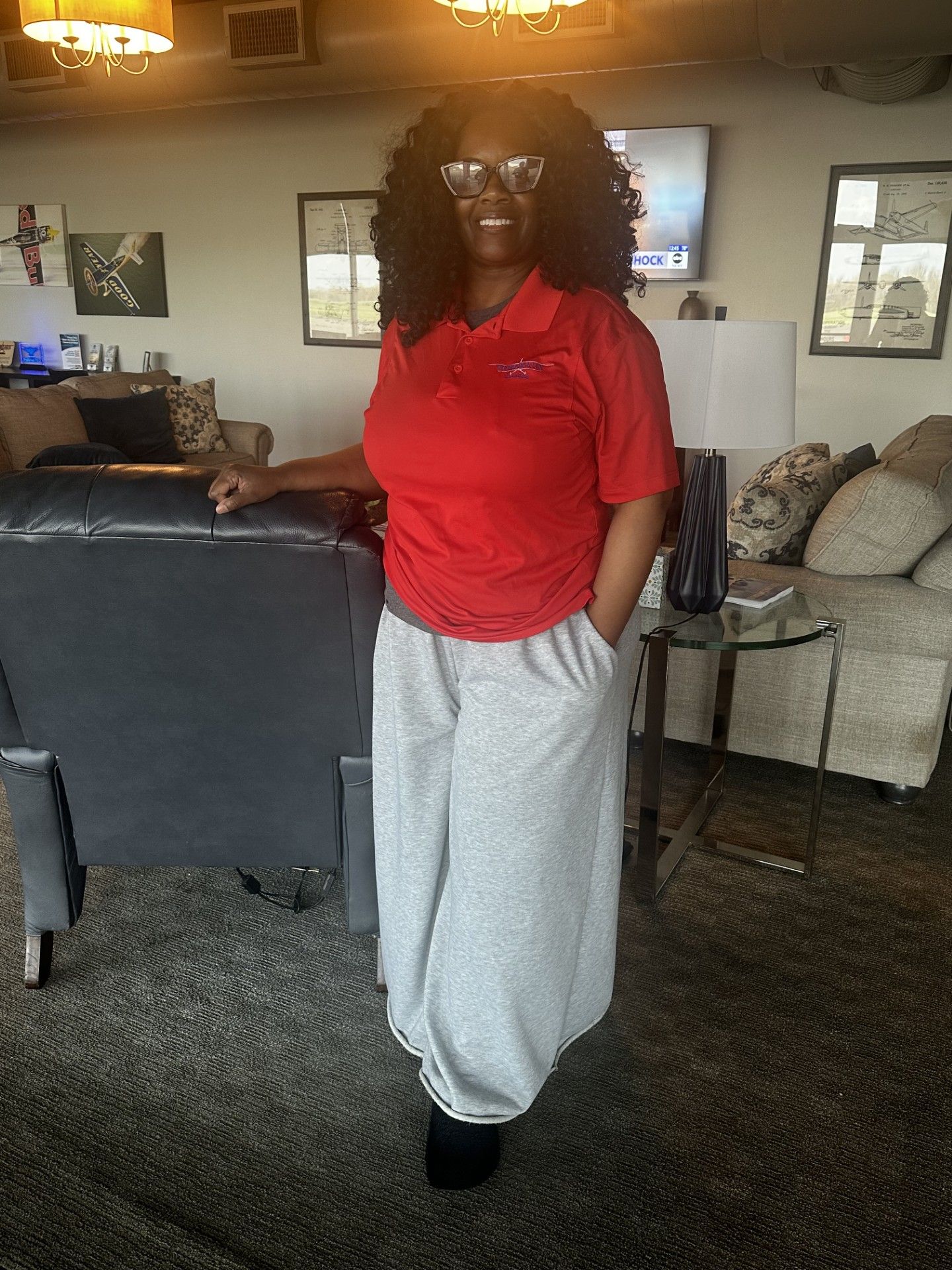 Person standing in a living room, wearing a red shirt and gray pants, beside a couch.