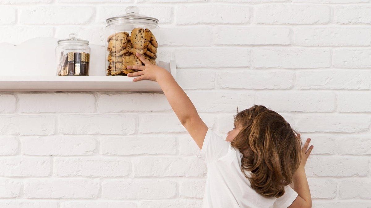 A little girl is reaching for a jar of cookies on a shelf.