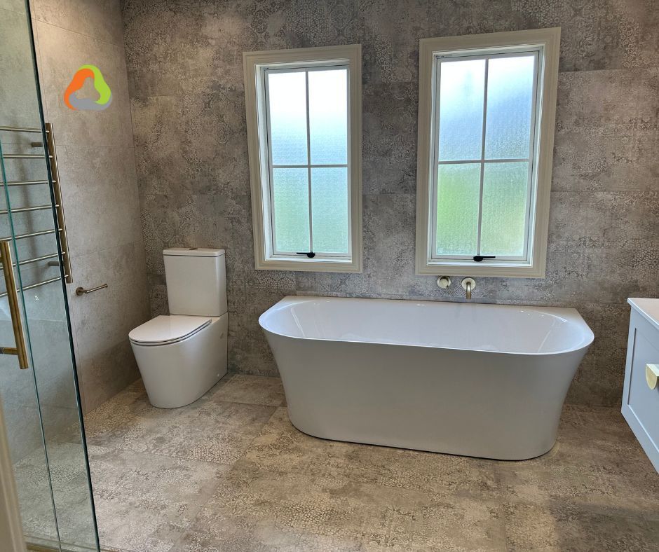 Modern bathroom with freestanding tub, toilet, glass shower, and two windows on tiled walls