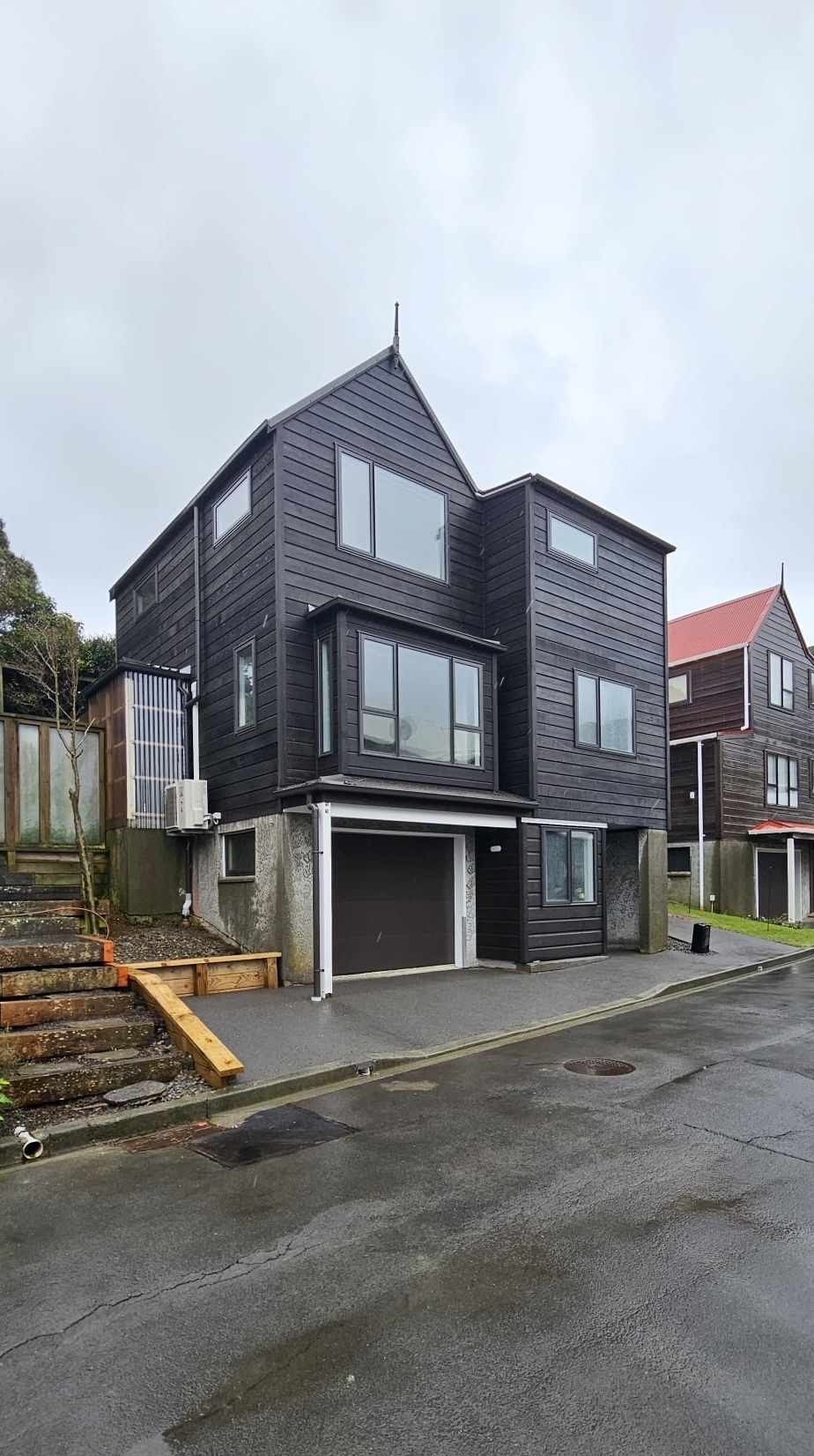 Modern dark-gray multi-story house with balconies on a wet street, viewed from the front