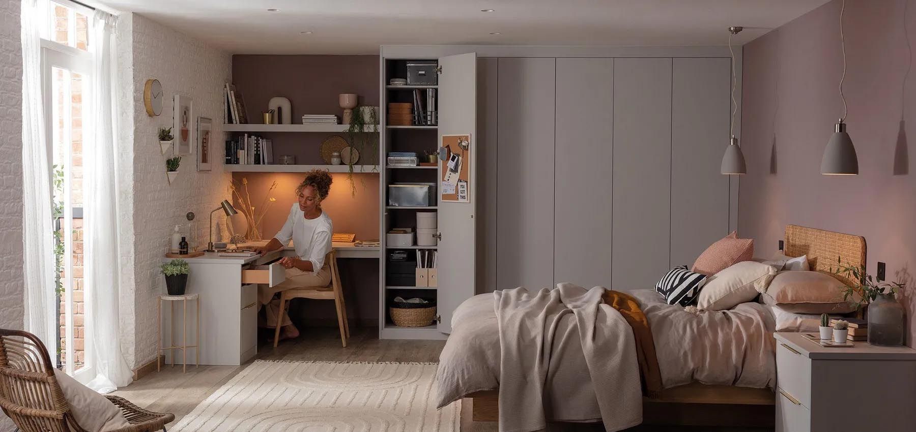 A woman is sitting at a desk in a bedroom next to a bed.