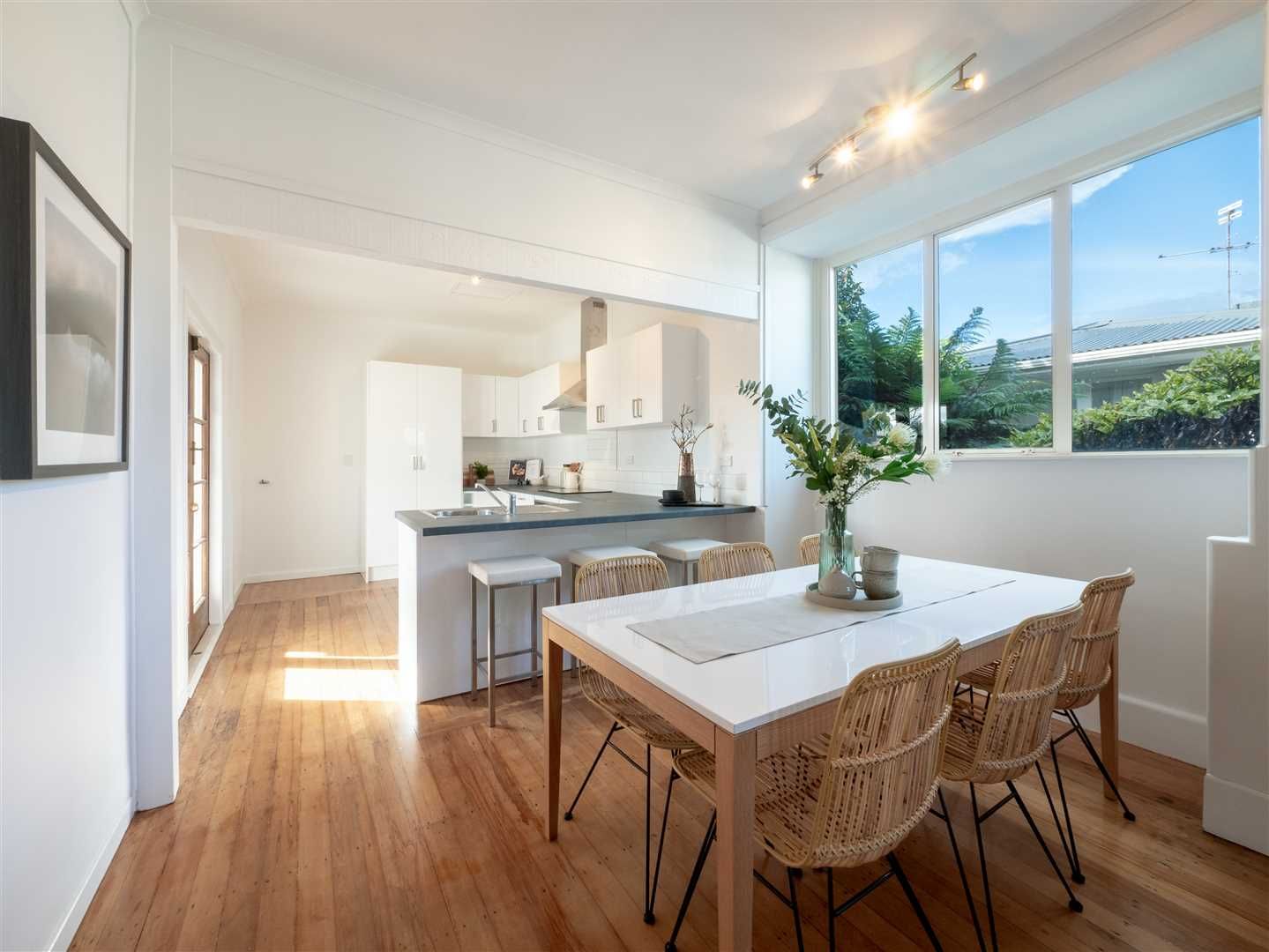 Bright open dining area with white walls, wood floors, wicker chairs, and large windows overlooking greenery