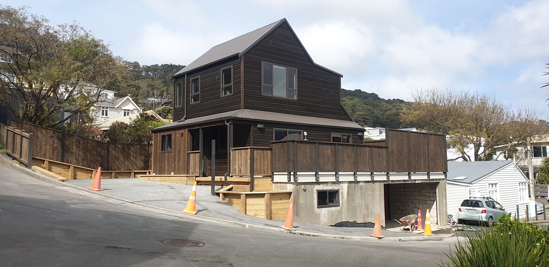 Two-story dark wood building under construction beside a sloped road with orange cones and a white truck