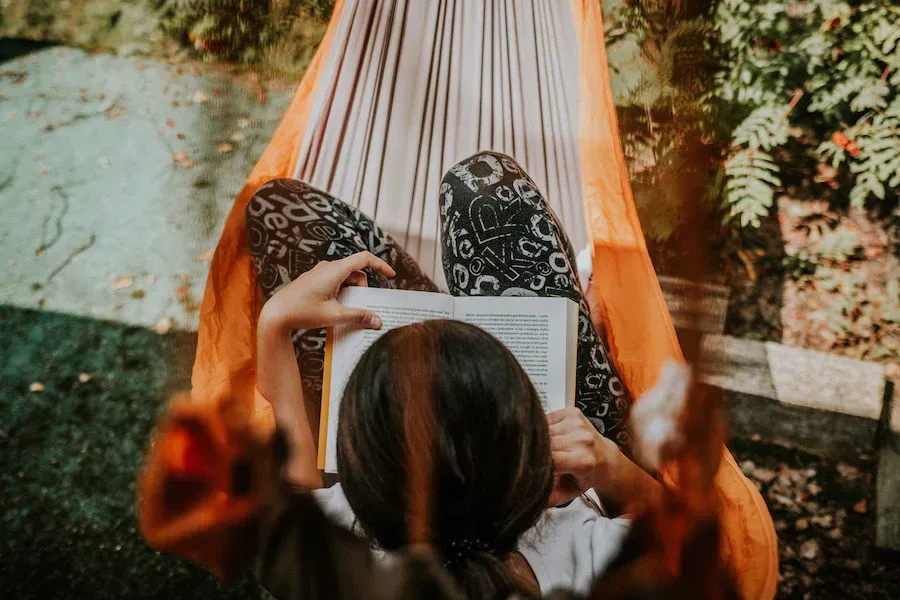 A woman is laying in a hammock reading a book.
