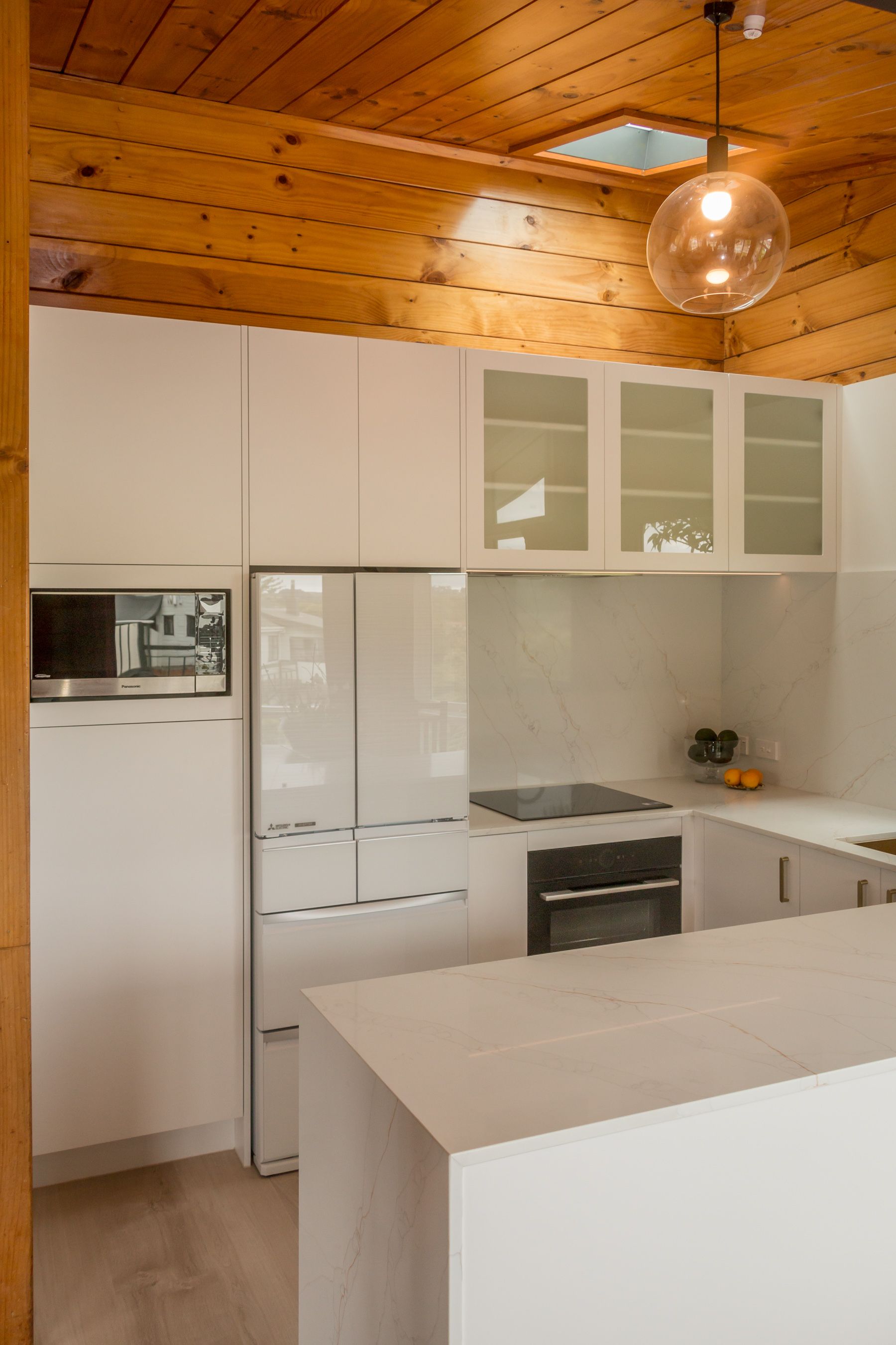 Modern white kitchen with wood-paneled ceiling, island, glass cabinets, and stainless appliances