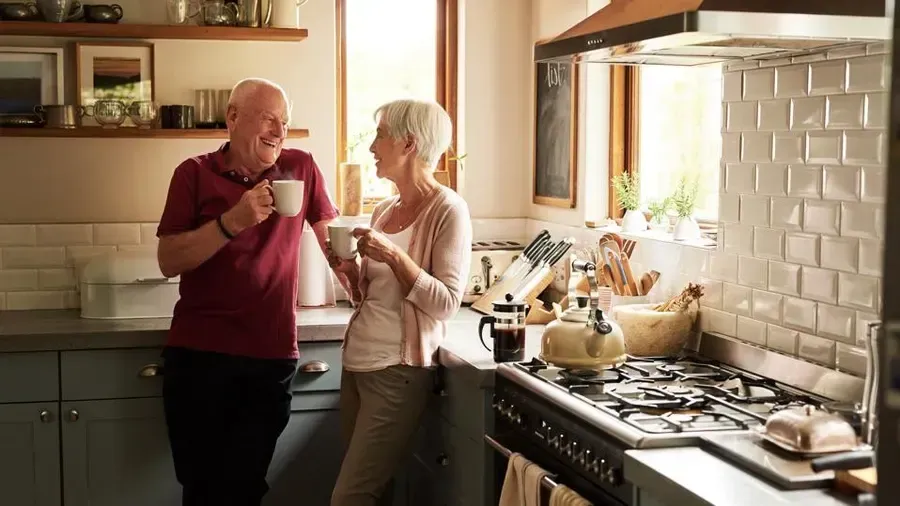 An elderly couple is standing in a kitchen drinking coffee.