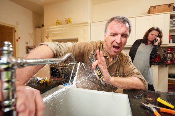 A man is fixing a sink in a kitchen while a woman talks on a cell phone.