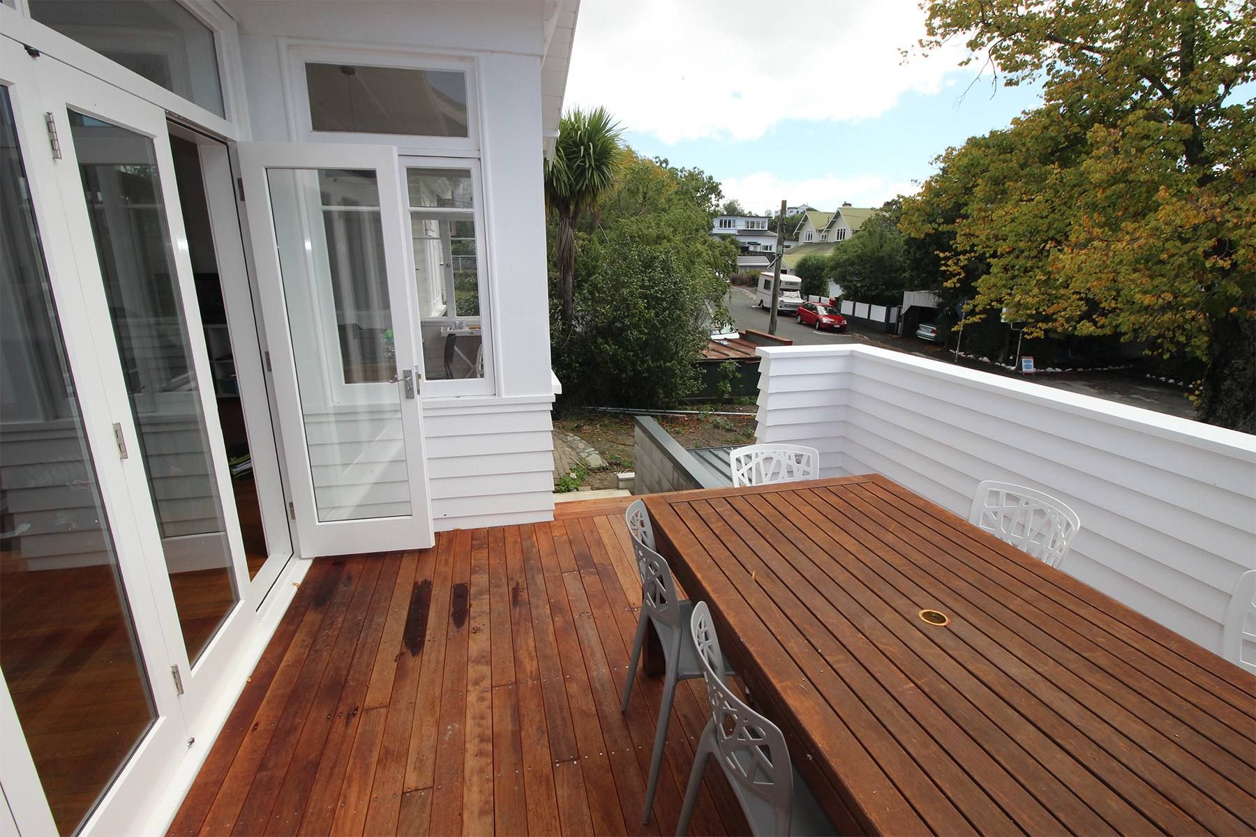 Wooden deck with white railing beside a house, overlooking trees and a street.