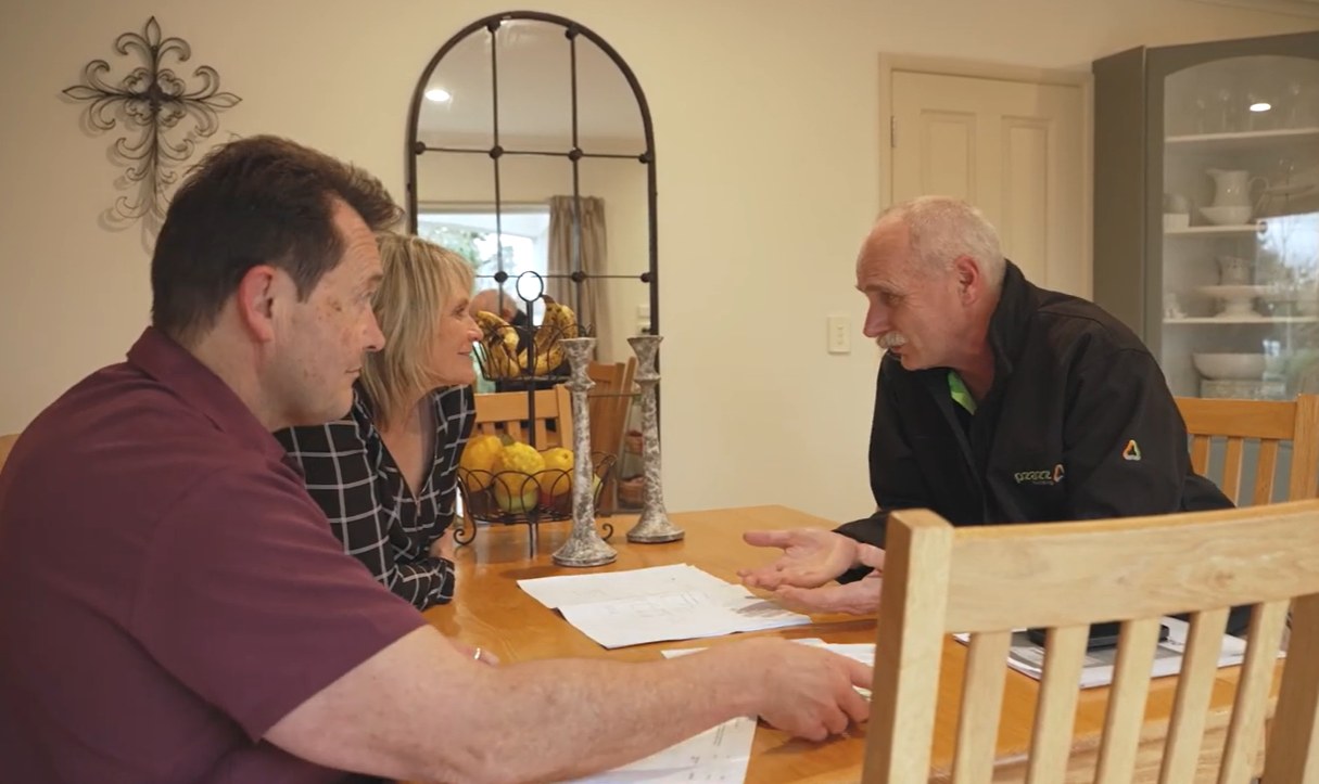 Three people sit at a dining table in a home, talking with papers spread out between them.