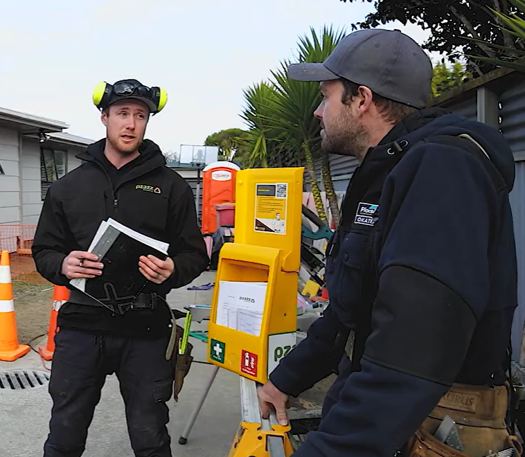 Two workers discuss a yellow equipment station outdoors, one holding papers and the other leaning on it.