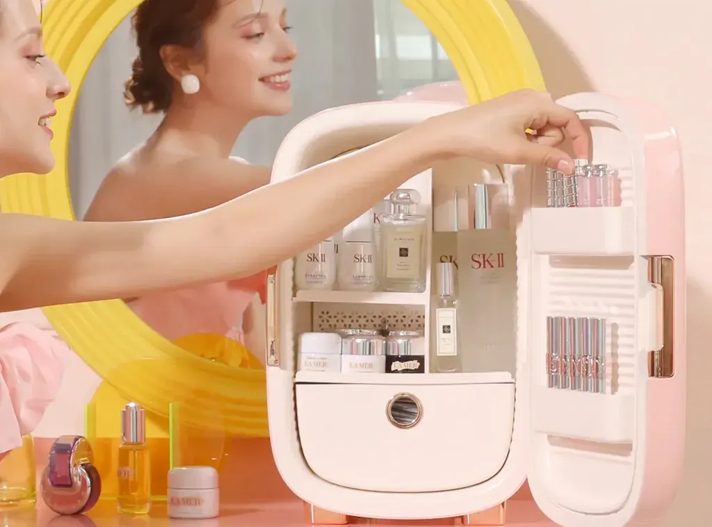 A woman is looking at her reflection in a mirror while opening a small fridge filled with cosmetics.