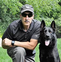 Man kneels beside black German Shepherd outdoors, both facing the camera.