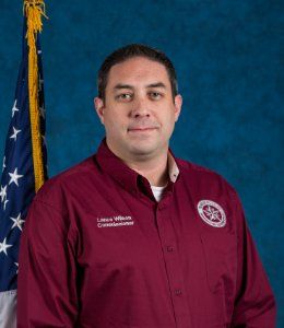 A man in a maroon shirt is standing in front of an american flag.