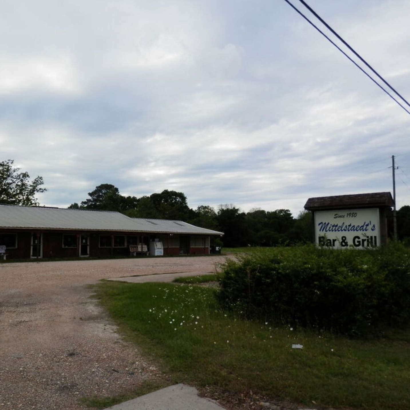 Rural roadside bar and grill with a sign, gravel lot, and cloudy sky