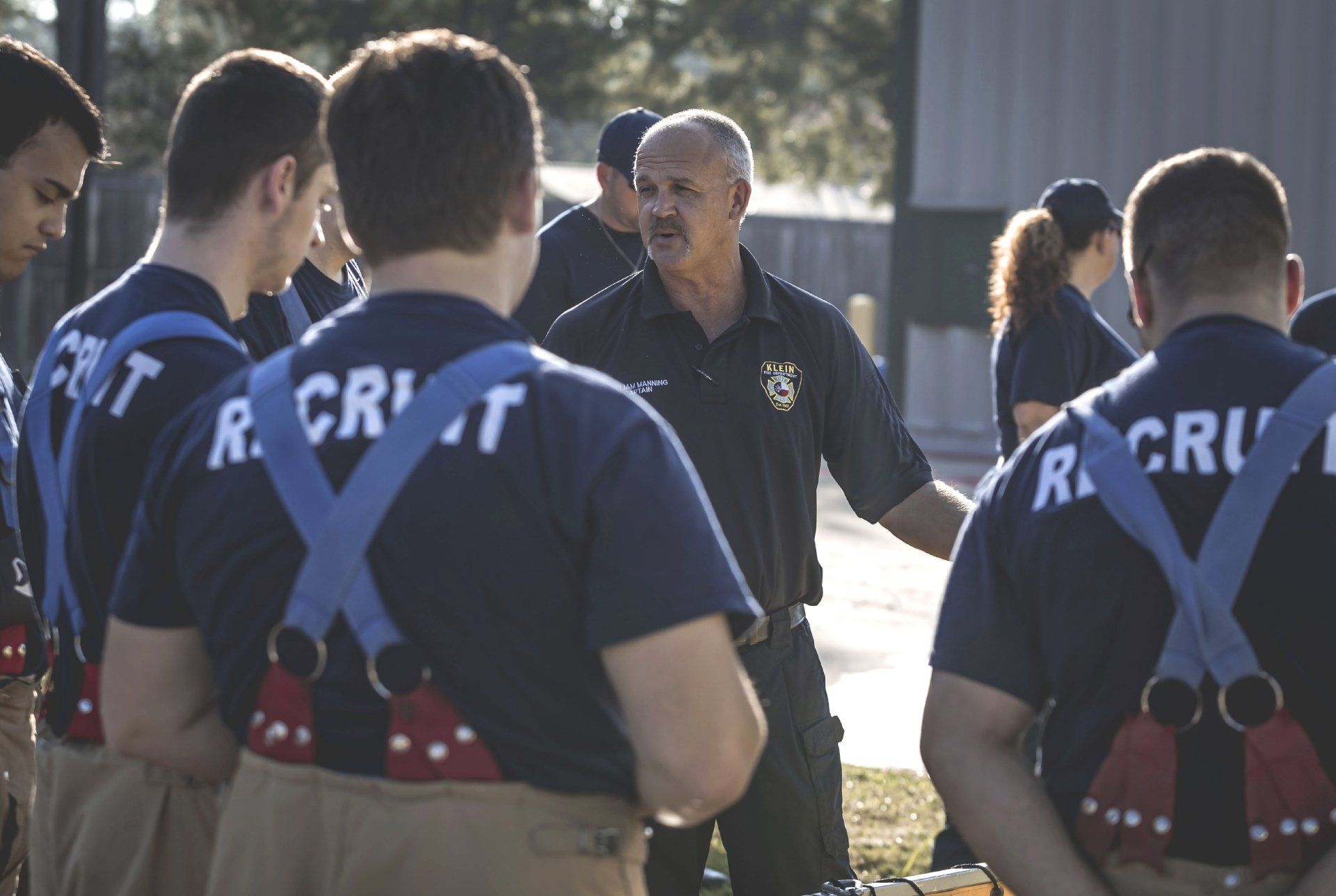 A man is talking to a group of firefighters with recruit on their shirts
