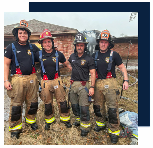 Four firefighters in turnout gear standing together outdoors in front of a brick building