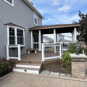 A wood-tone deck with white railings and a roofed section attached to a grey-sided house, next to a stone pillar.