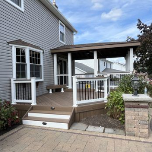 A wood-tone deck with white railings and a roofed section attached to a grey-sided house, next to a stone pillar.