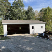 A white, single-story garage with a gray shingled roof, featuring an open double bay and two windows, set in a wooded area.