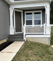 A grey house with a covered porch, white columns and railing, grey stairs, and a dark brown front door.
