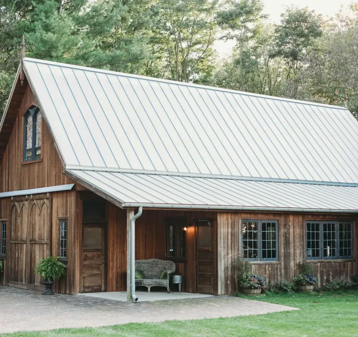 A rustic wooden barn with a metal roof and a covered porch, set amidst lush green trees and foliage.