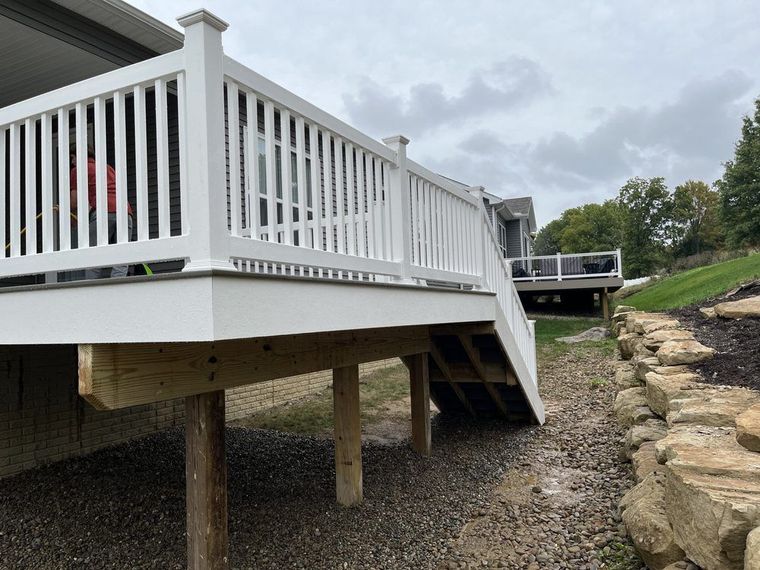 A low-angle view of a white deck with a railing, supported by wooden posts above a gravel ground and a stone retaining wall.