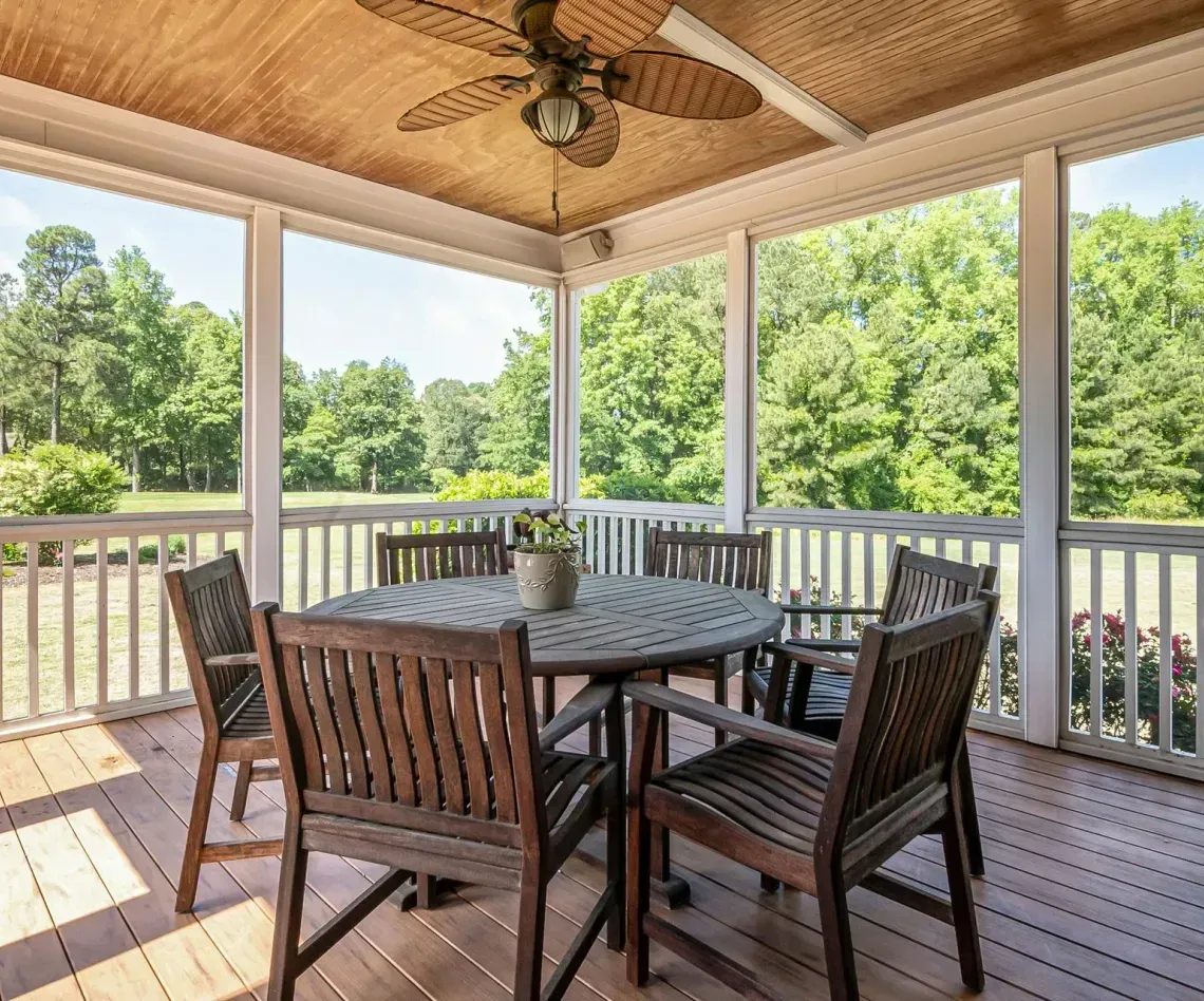 A wooden porch with a round table and chairs overlooking a green, tree-filled landscape.