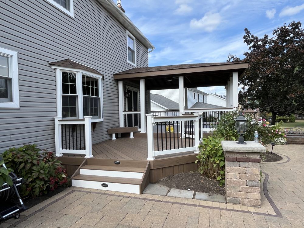 A gray house exterior with a brown wooden deck, white railings, a covered patio area, and a brick paver walkway.