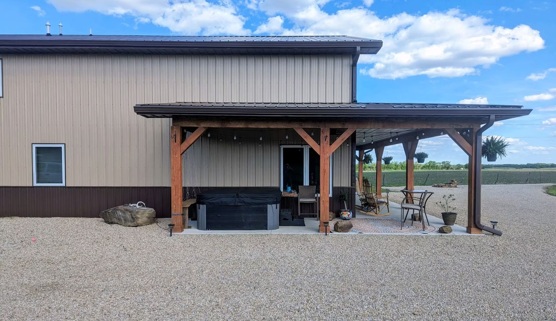 A two-story beige and brown metal building with a brown wooden pergola, seating, and gravel yard.