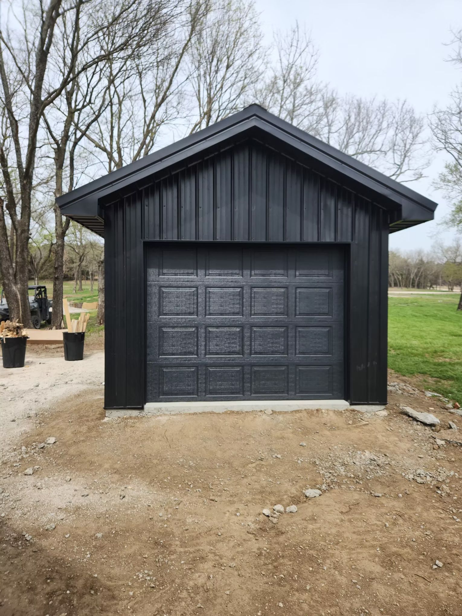 Black garage with a matching garage door, set on a dirt lot, with trees in the background.