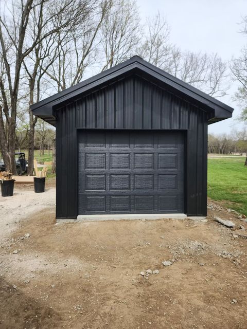 Black metal garage with a matching door and roof against a backdrop of trees and a dirt lot.