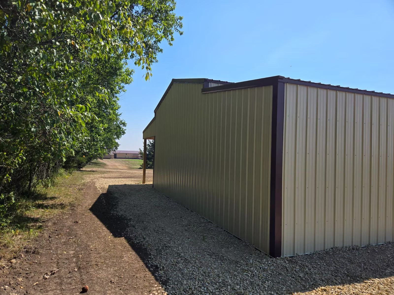 Tan metal building with brown trim, gravel foundation, and dirt path alongside green trees on a sunny day.