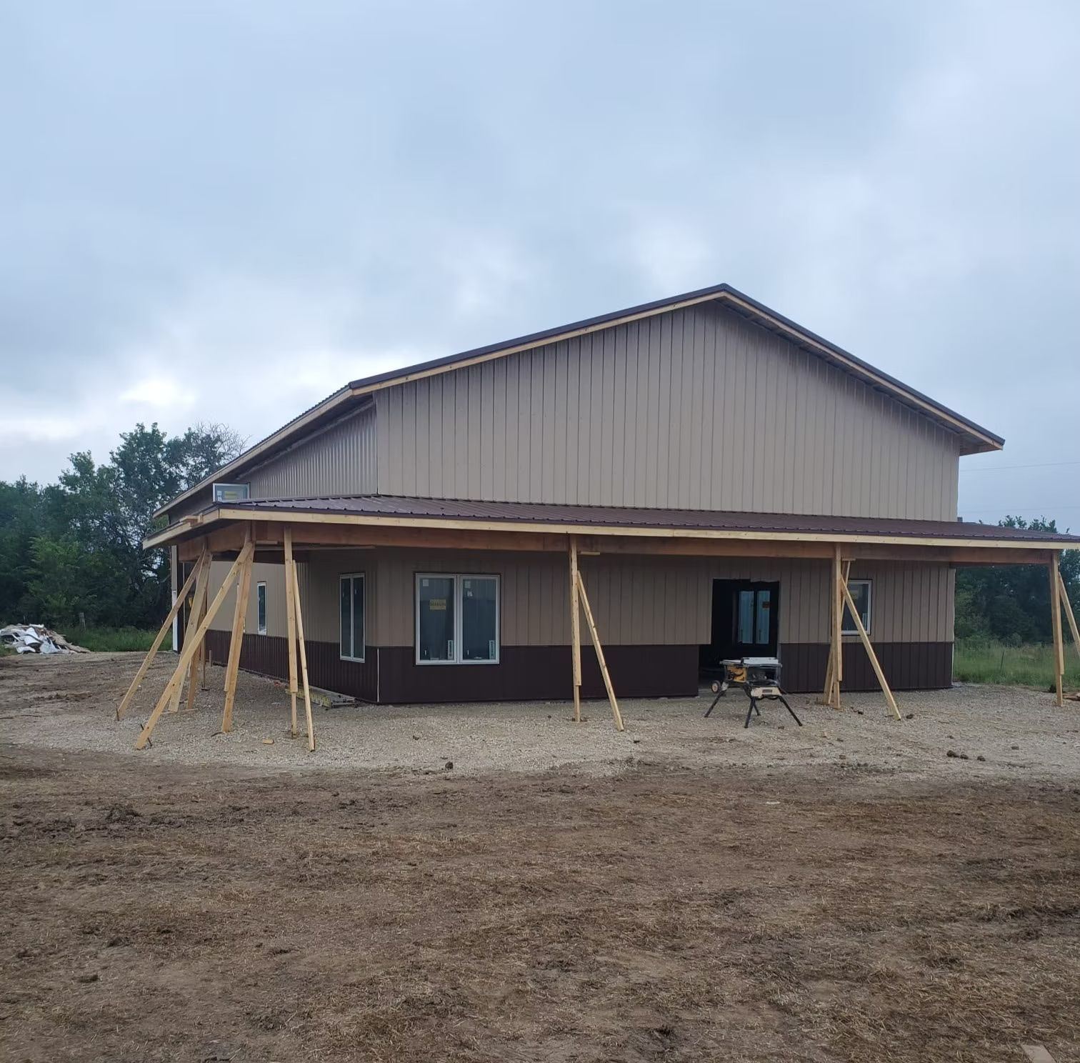 A two-story building under construction, with tan and brown  a covered porch supported by wooden posts, set on a gravel lot.
