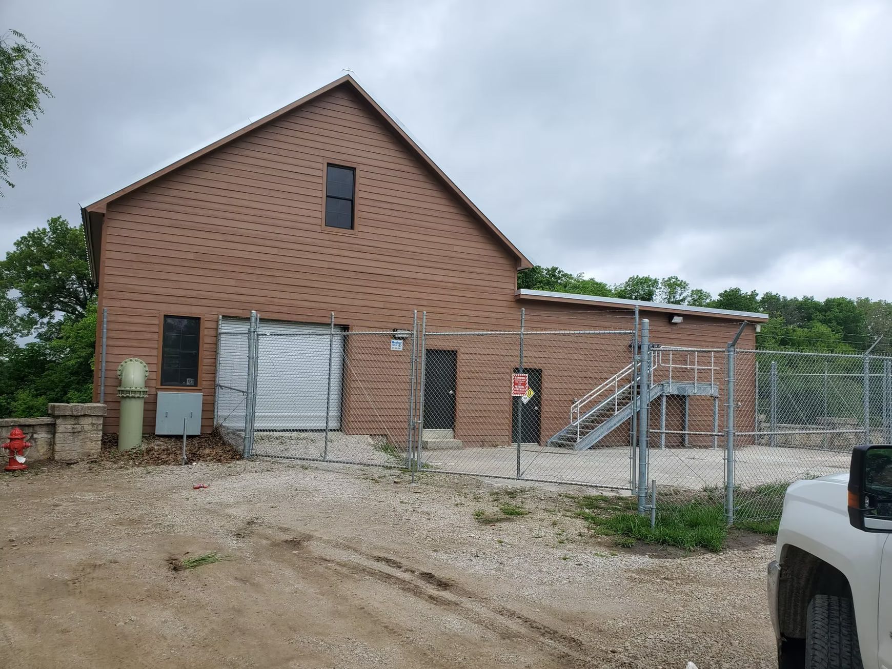 Brick building behind a chain-link fence, on gravel, under a cloudy sky. A white truck is partially visible.