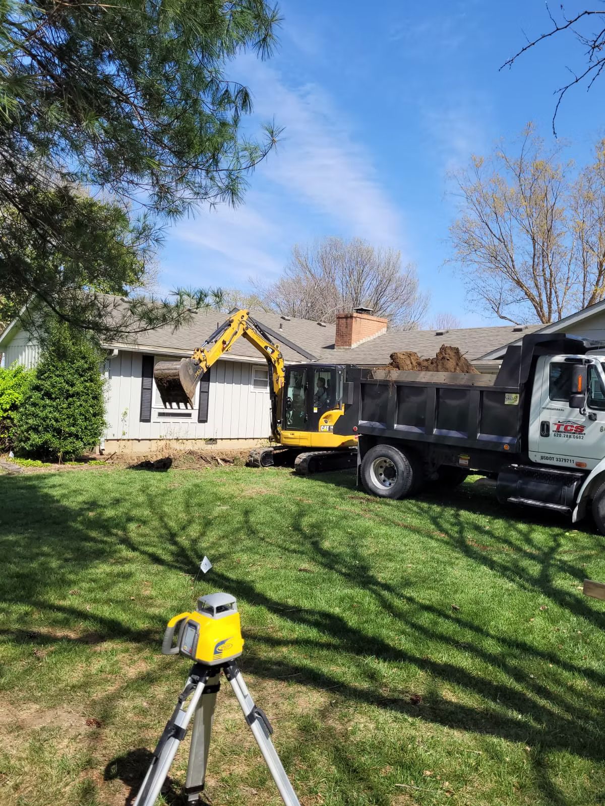 Excavator digging near a house, loading a dump truck. A laser level sits in the foreground on the grass, under a blue sky.