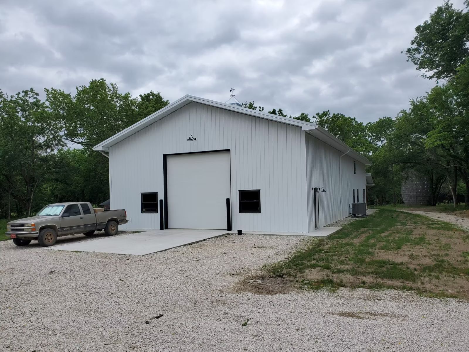 White metal building with a large overhead door, small windows, and a gray pickup truck parked in front on a gravel driveway.
