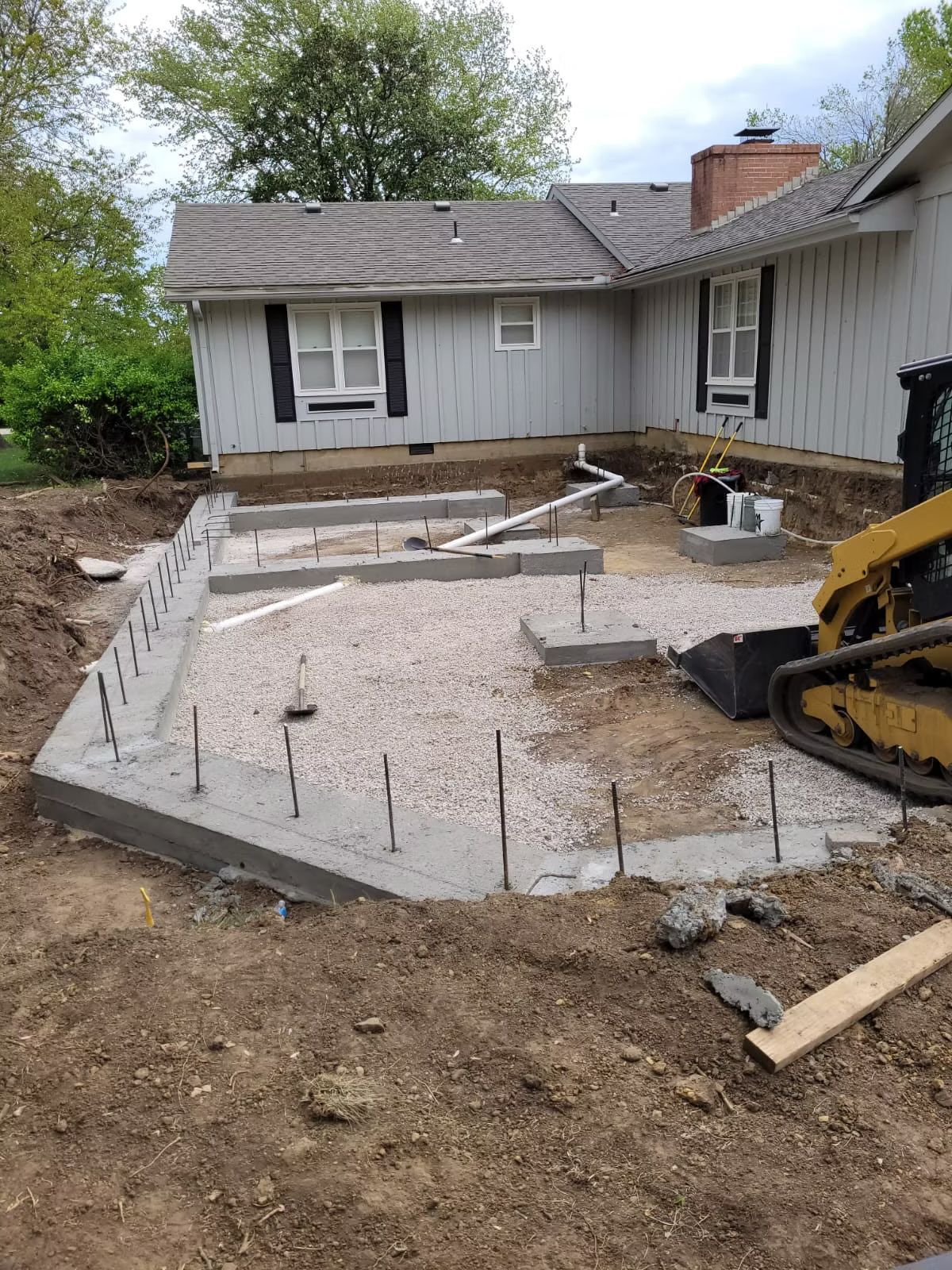 A concrete patio foundation under construction next to a house. Gray concrete forms the edge, with gravel and rebar inside.