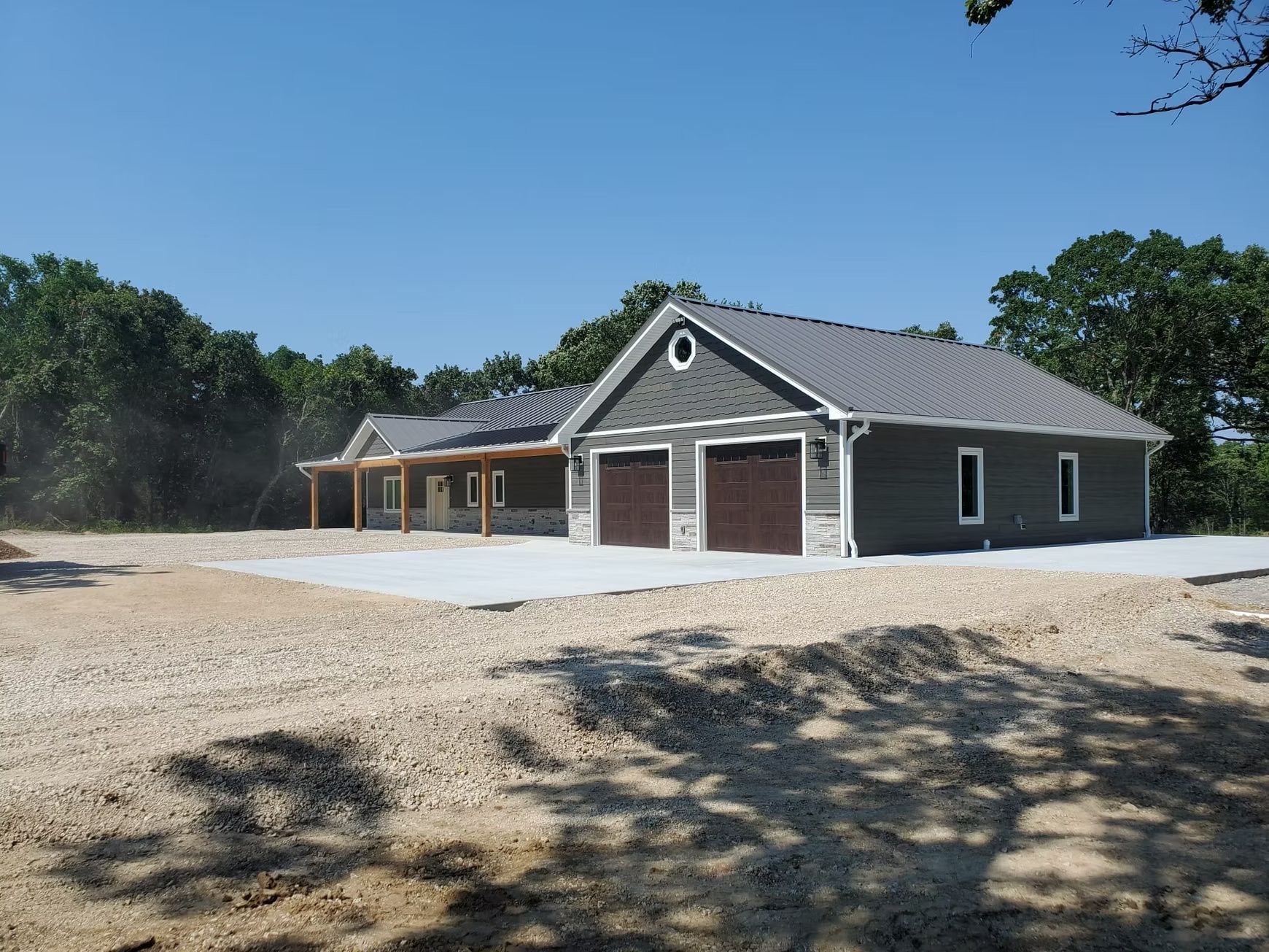 A newly built, single-story house and garage with a gravel driveway under a bright blue sky, surrounded by trees.