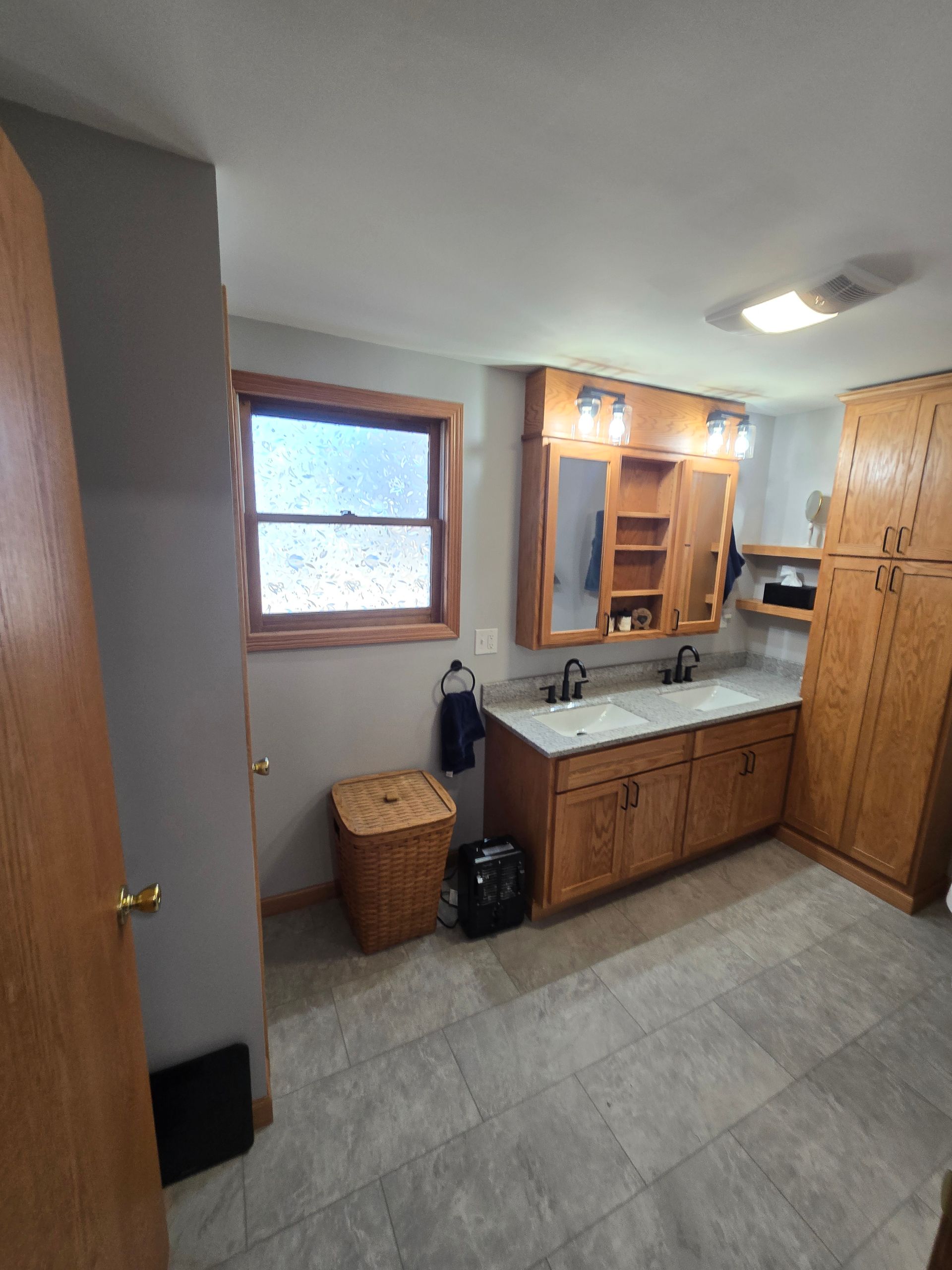 A bathroom with a double vanity, wooden cabinetry, tiled floors, a window, and a wicker hamper.