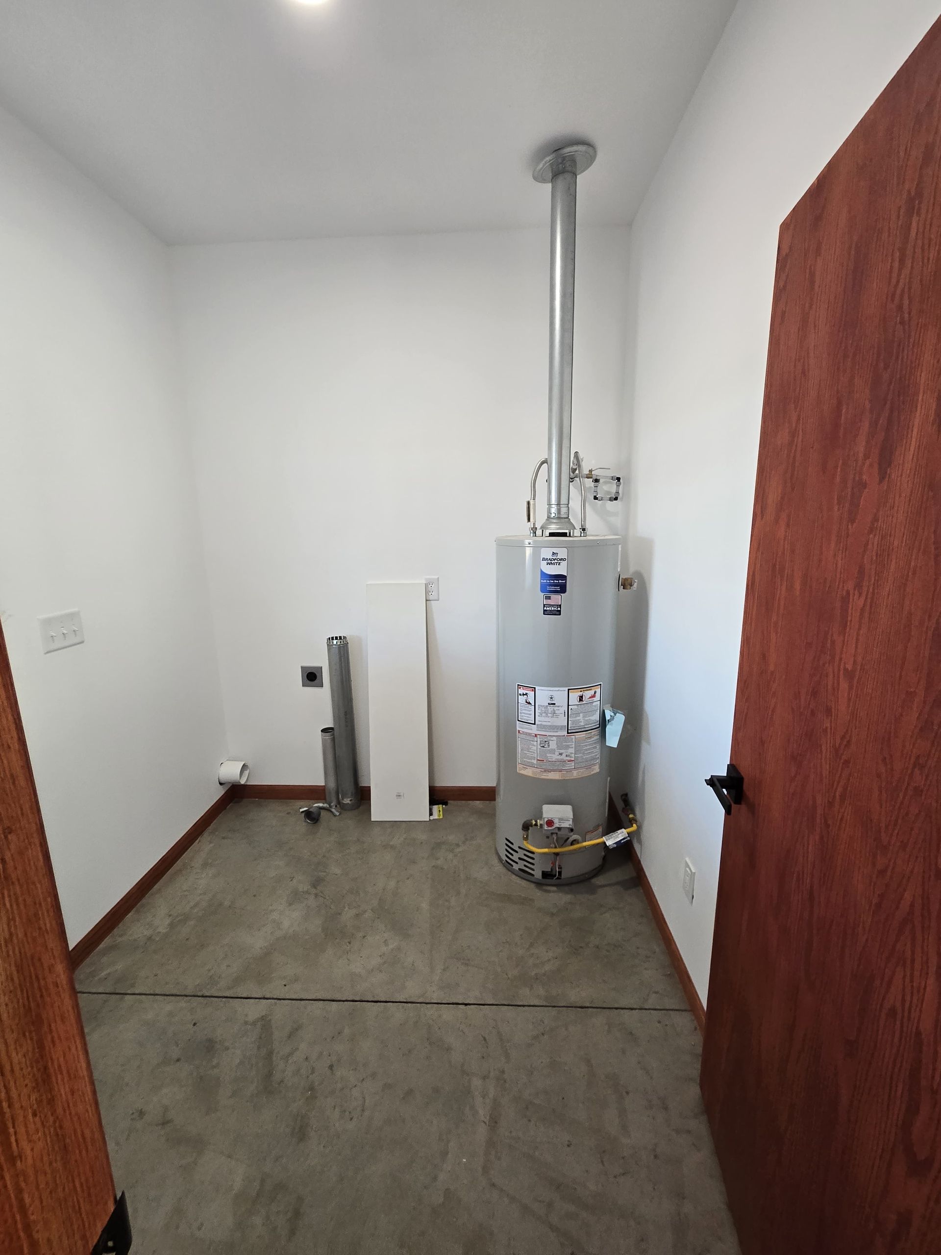 Utility closet with a water heater, metal exhaust pipe, and concrete floor, viewed through an open wooden door.