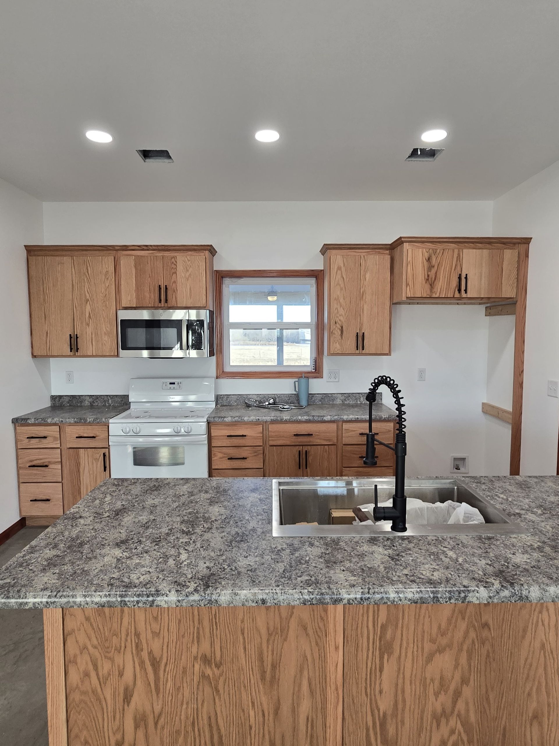 A kitchen with wood cabinets, gray speckled countertops, an island with a black faucet, and white appliances.