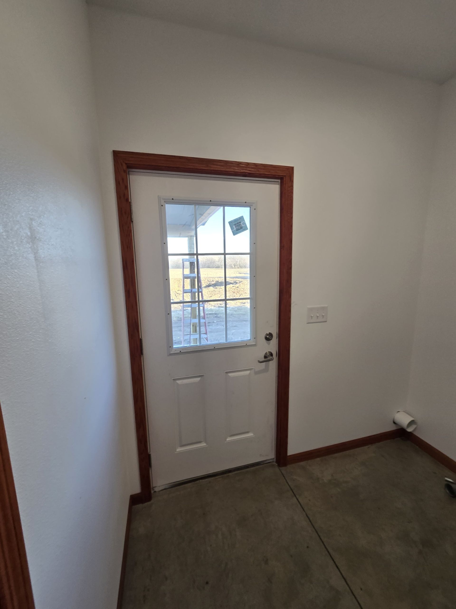 White door with a glass window and brown trim in a room with white walls and a concrete floor.